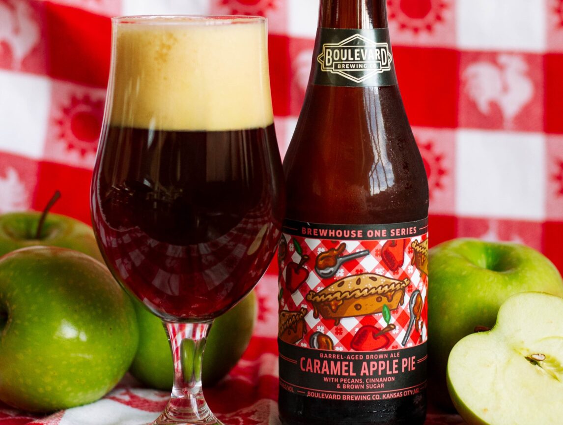 A bottle of Boulevard Brewing Co. Caramel Apple Pie beer and a glass of dark beer are placed on a red and white checkered cloth, surrounded by whole and sliced green apples.