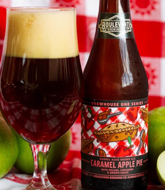 A bottle of Boulevard Brewing Co. Caramel Apple Pie beer and a glass of dark beer are placed on a red and white checkered cloth, surrounded by whole and sliced green apples.