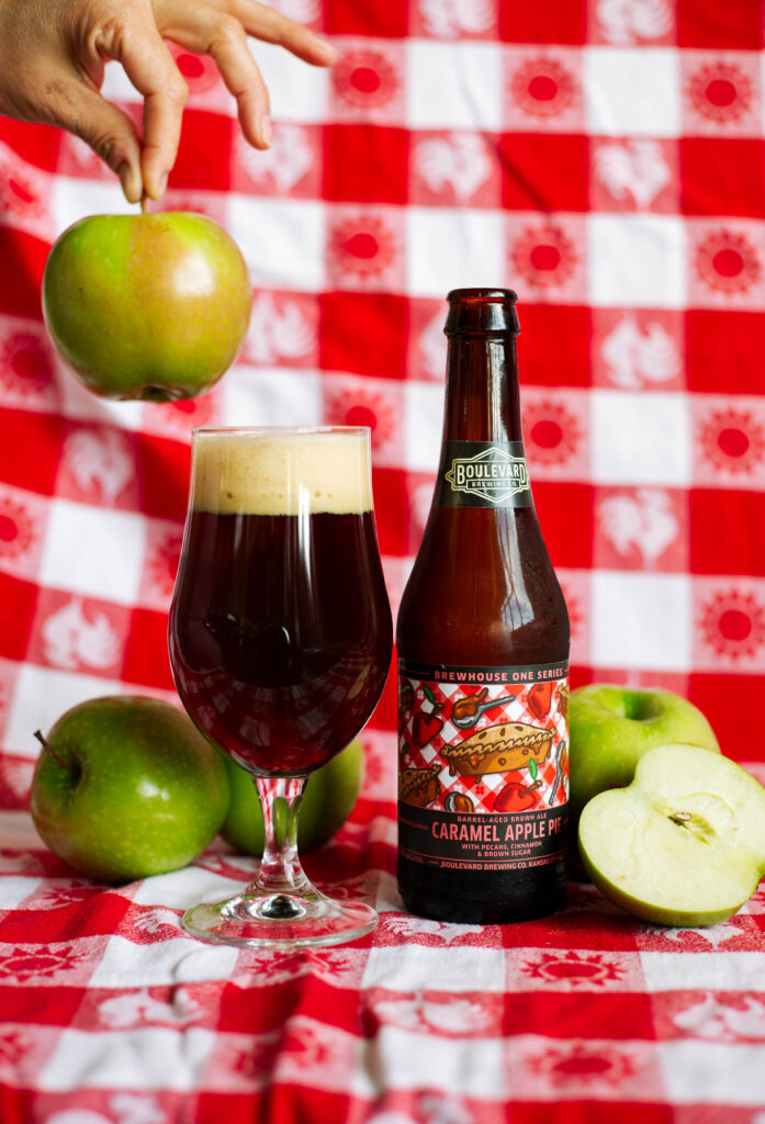 A bottle of Boulevard Caramel Apple Pie beer stands next to a glass of dark beer with foam. Green apples, a halved apple, and a hand holding an apple appear on a red and white checkered tablecloth background.