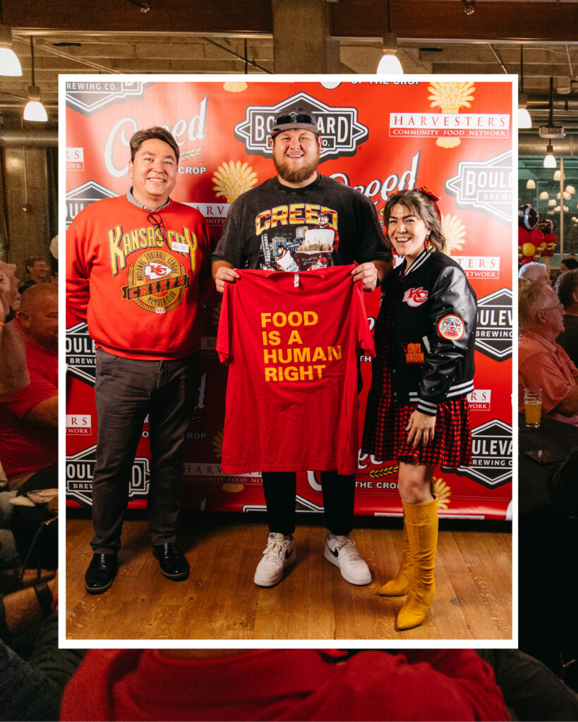 Three people pose and smile at an event. The person in the middle, standing proudly with Creed Humphrey, holds a red shirt reading “FOOD IS A HUMAN RIGHT.” The background features Boulevard-branded banners and a crowd sitting at tables.