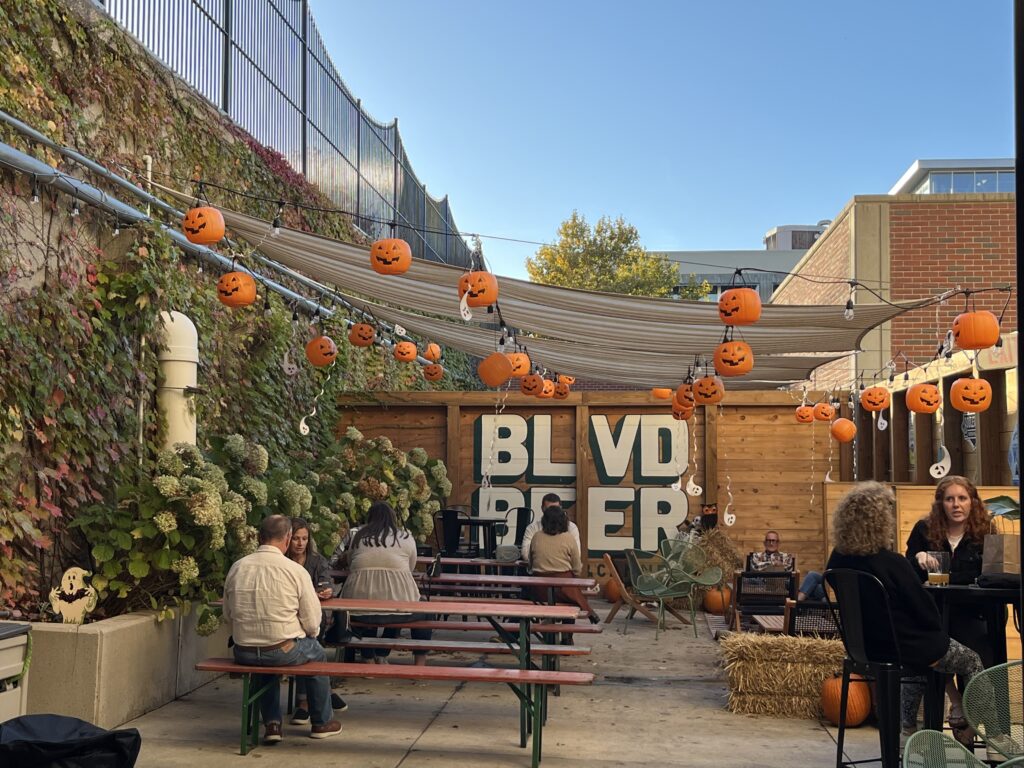 People sit at picnic tables in an outdoor patio decorated with hanging jack-o'-lantern lanterns for Rec Deck the Halls. Large "BLVD BEER" letters, autumn plants, and hay bales enhance the festive atmosphere.