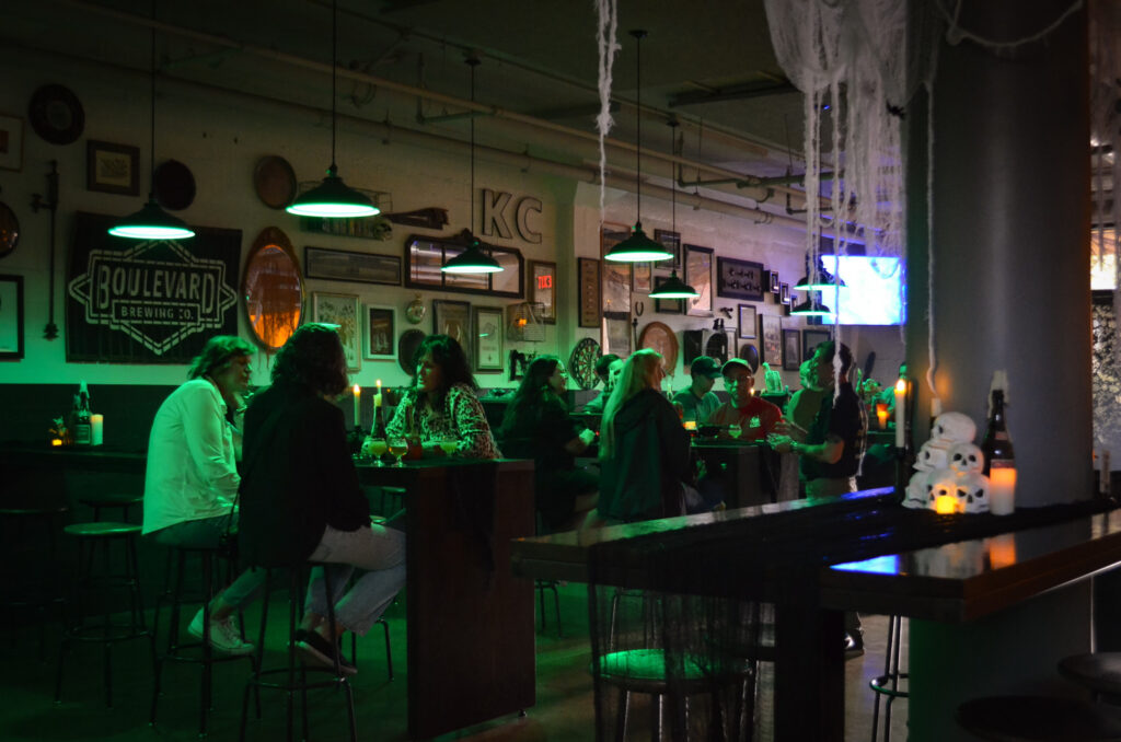 A group of people sit and talk at tables in the cozy, casual bar, decorated with green lights, Halloween decorations, and wall art, creating a warmly festive atmosphere.