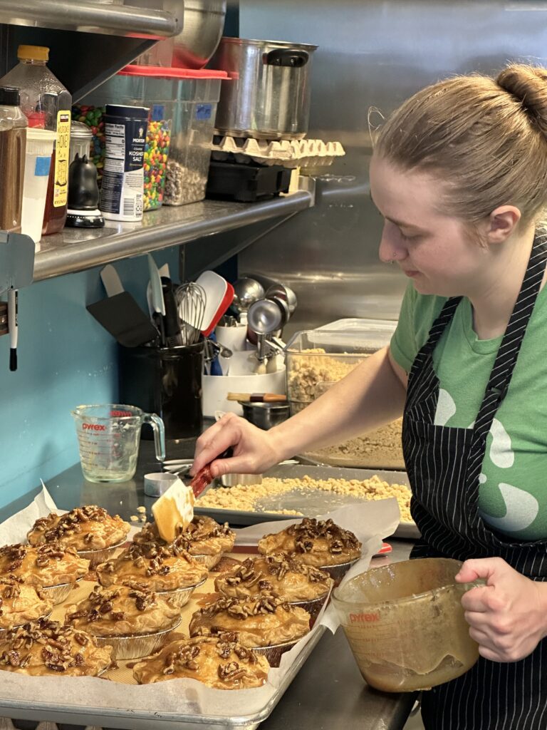 A woman in a striped apron spreads a topping onto trays of cinnamon rolls with pecans in a kitchen, surrounded by baking tools and ingredients.