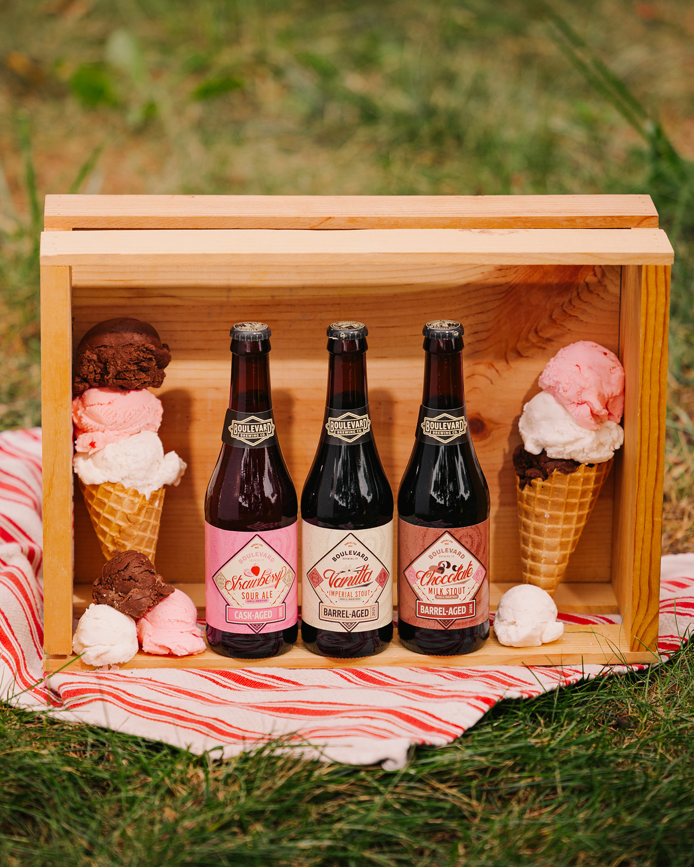 Three bottles of barrel-aged beer are displayed in a wooden crate, surrounded by waffle cones filled with scoops of pink and chocolate ice cream, set on a red-and-white striped cloth on grass.