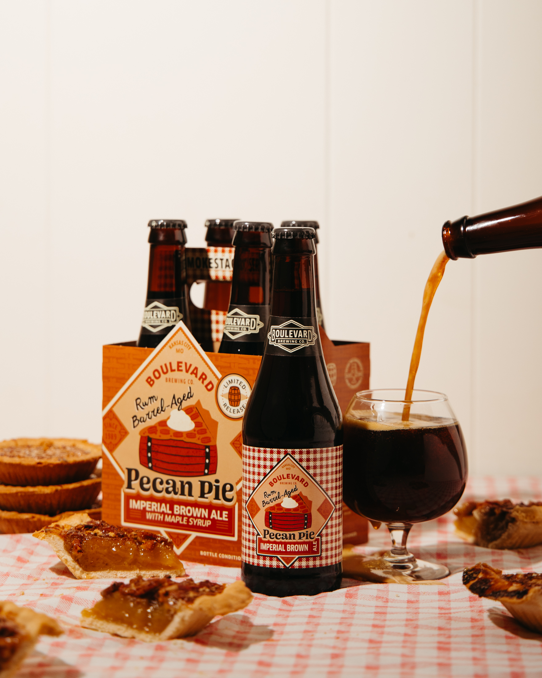 A glass of dark beer being poured next to a bottle and a six-pack of Boulevard Pecan Pie Imperial Brown Ale, with slices of pecan pie on a red checkered tablecloth.