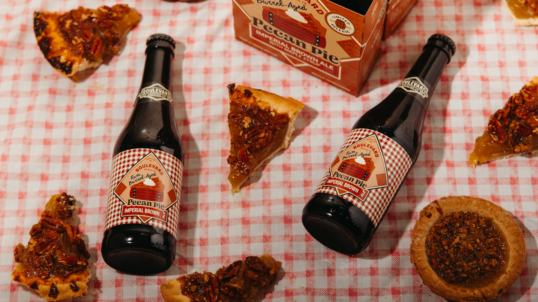 Two bottles of pecan pie imperial brown ale on a red and white checkered cloth, surrounded by slices of pecan pie and a pecan tart. A six-pack box of the beer is partially visible in the background.