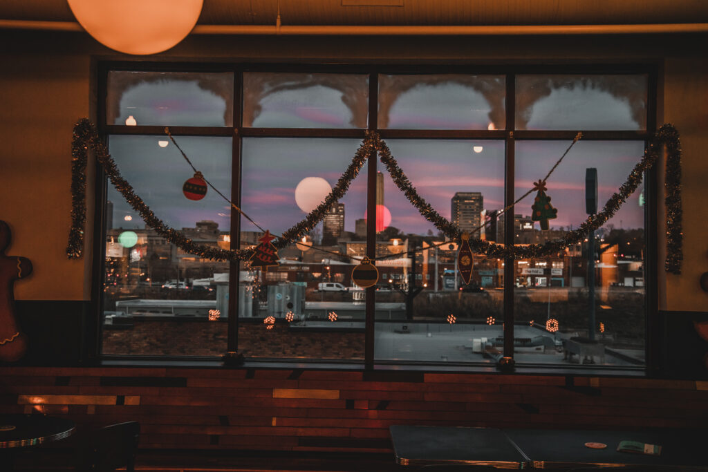 A decorated window with tinsel and holiday ornaments, channeling Rec Deck the Halls vibes, overlooks a cityscape at dusk. Buildings and a purple-pink sunset glow in the background while indoor lights reflect softly on the glass.