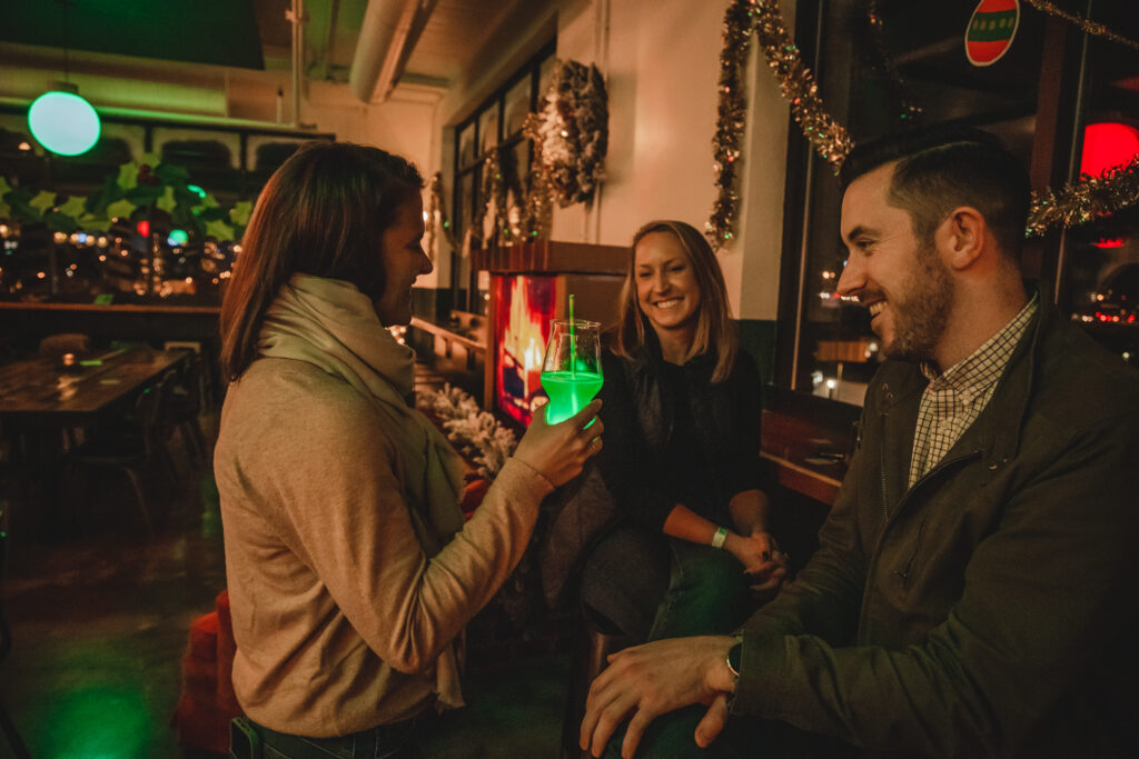 Three people are socializing in a festive, warmly lit room decorated with garlands. One woman holds a glowing green drink while talking to a man and another woman, sharing laughs as they Rec Deck the Halls together.