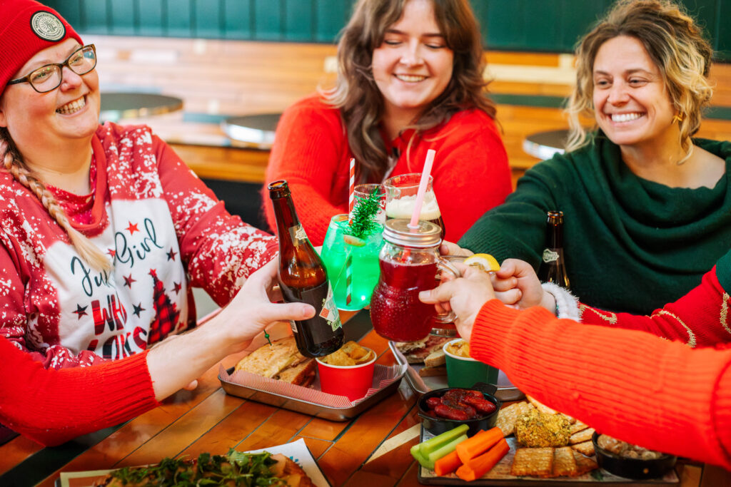 Four smiling women in festive clothing raise drinks together over a table filled with snacks, pizza, and dips, enjoying a cheerful Rec Deck the Halls gathering at a lively restaurant.
