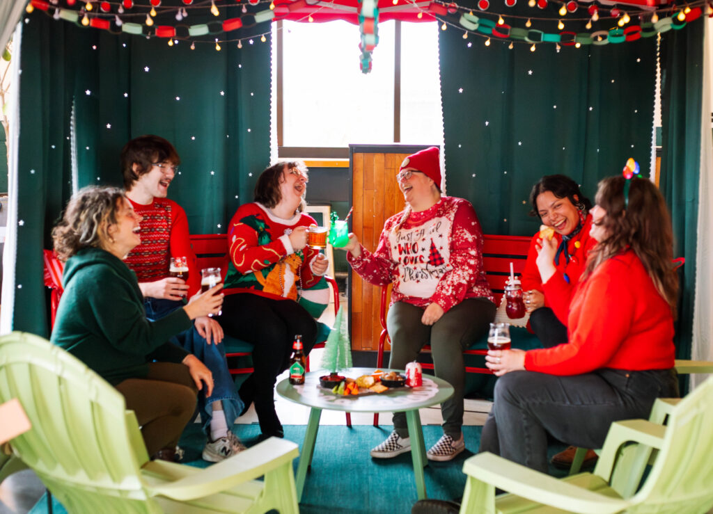 Six people in festive sweaters laugh and toast with drinks around a small table of snacks, enjoying a cozy Rec Deck the Halls gathering surrounded by string lights and holiday decorations.