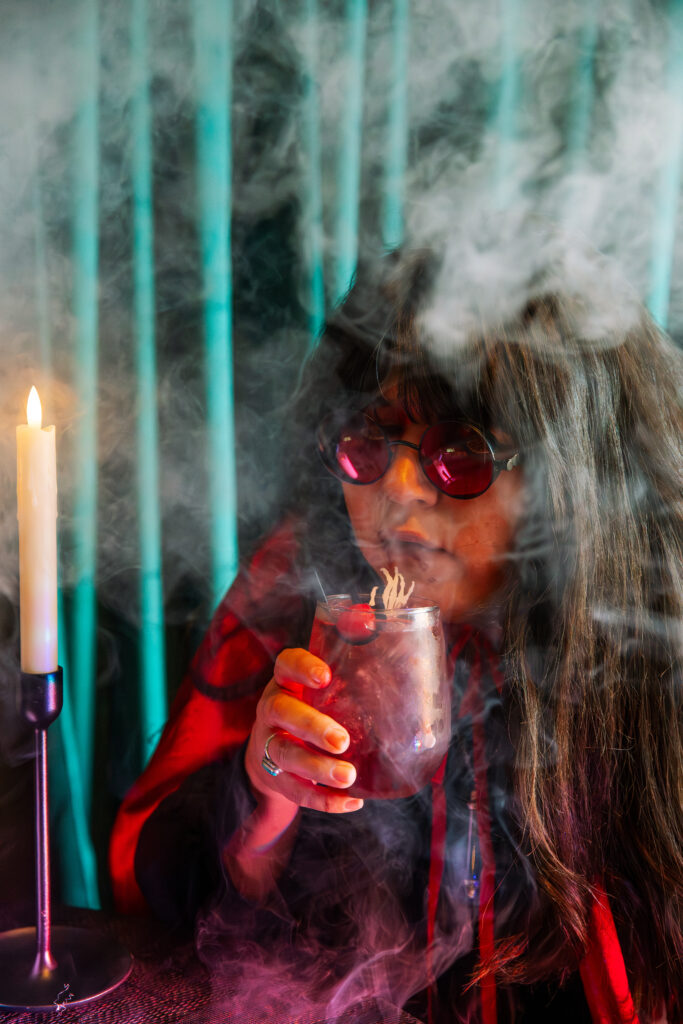A person with long hair and red sunglasses holds a smoky drink near their face, surrounded by thick mist at Boolevard. They wear a red and black outfit, with a lit candle on the left and green curtains in the background.
