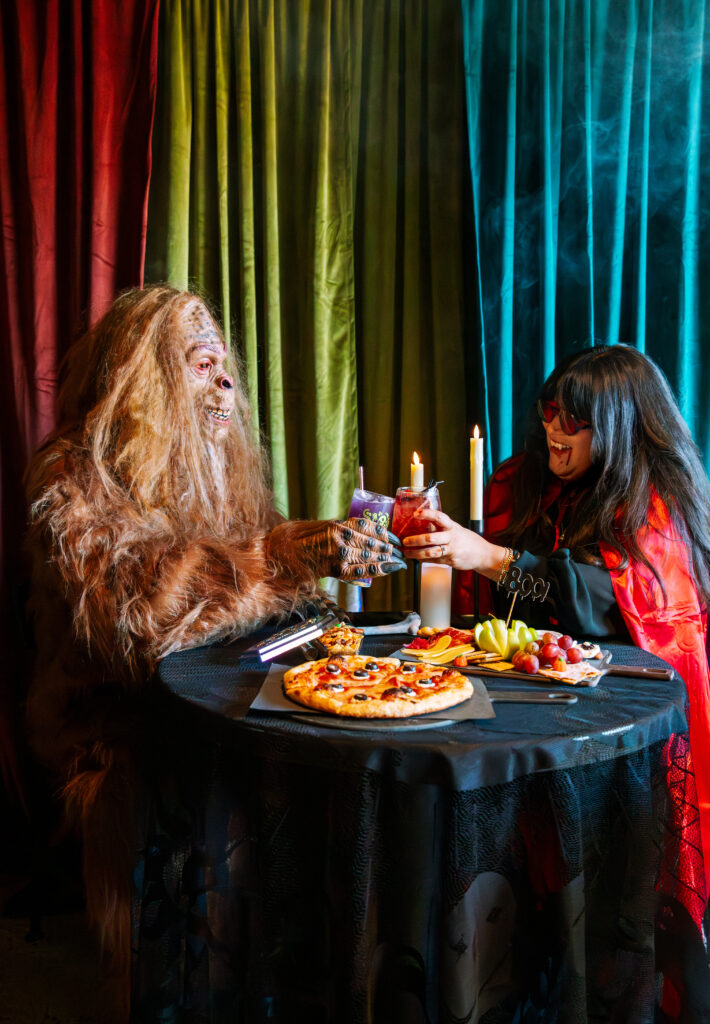 A person in a furry monster costume and another in a red cape and sunglasses clink glasses at a candlelit table with pizza and snacks at Boolevard, set against colorful curtains and a smoky, festive atmosphere.