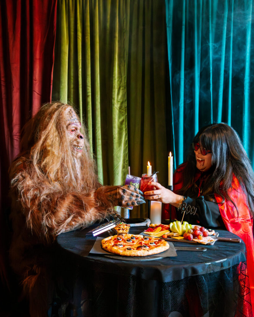 At Boolevard, a person in a Bigfoot costume and another in a witch costume toast drinks at a table with pizza, grapes, and candles, all set against colorful curtains.