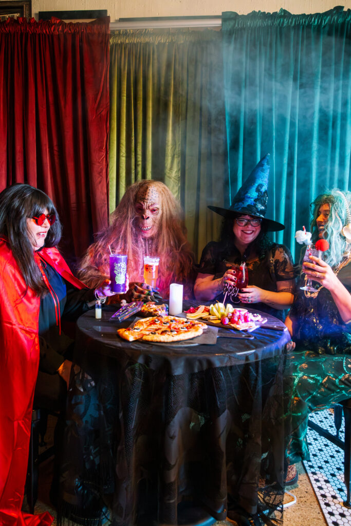 Four people in colorful Halloween costumes (vampire, hairy monster, witch, and sea witch) gather at a round table with pizza and drinks, laughing and celebrating Boolevard style in a festive, spooky setting with vibrant curtains and smoke.