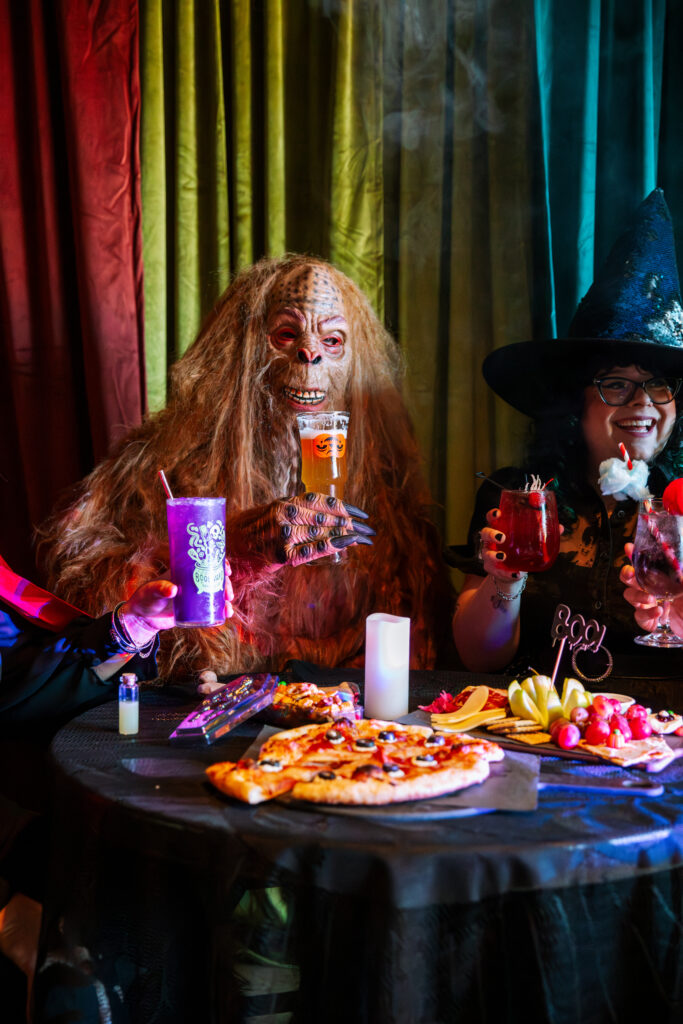 Two people in Boolevard Halloween costumes—a hairy monster and a witch—sit at a table with colorful drinks, pizza, and snacks, smiling together against vibrant curtains in a festive atmosphere.
