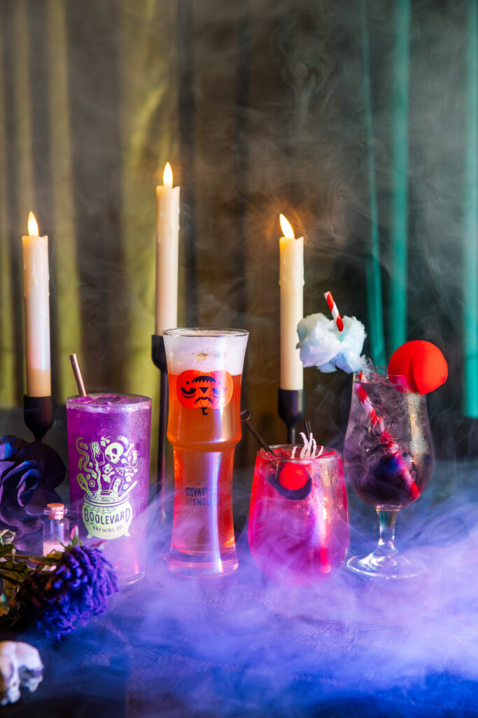 Four Boolevard cocktails, surrounded by flowers and dramatic fog, are displayed before four lit candles on a dark, moody background. Cotton candy, cherries, and unique garnishes crown the vibrant drinks.