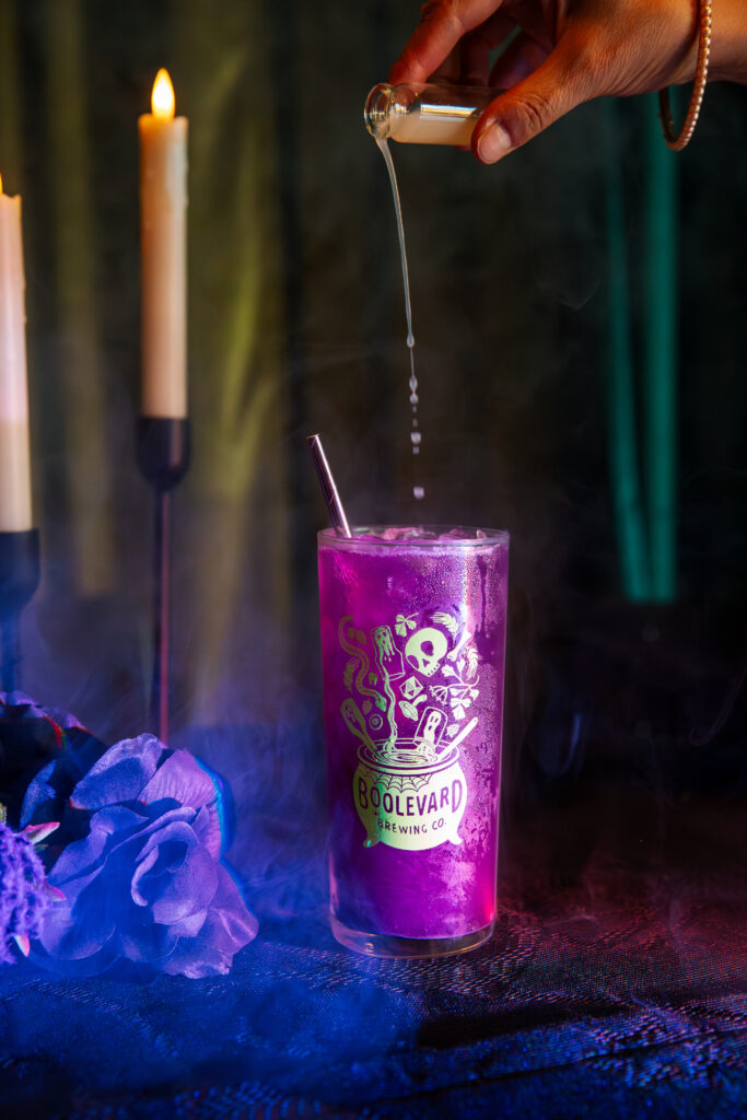 A vibrant purple Boolevard cocktail in a Boulevard Brewing Co. glass sits on a table with swirling smoke. A hand pours liquid from a vial into the drink, while two lit candles and blue flowers create a magical backdrop.