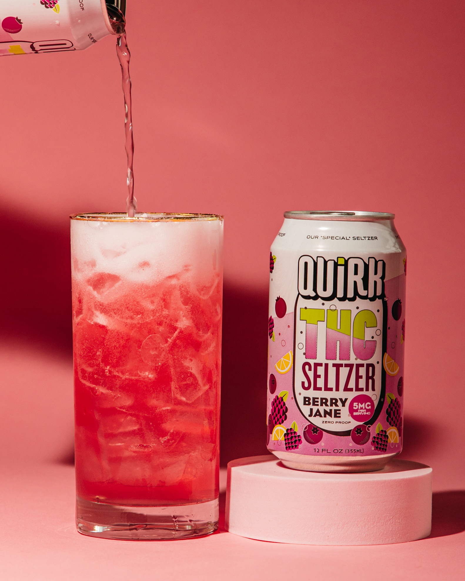 A can of Quirk THC Seltzer, Berry Jane flavor, sits beside a tall glass filled with ice and vibrant red THC Seltzer being poured in, set against a pink background. The can is adorned with colorful fruit illustrations.