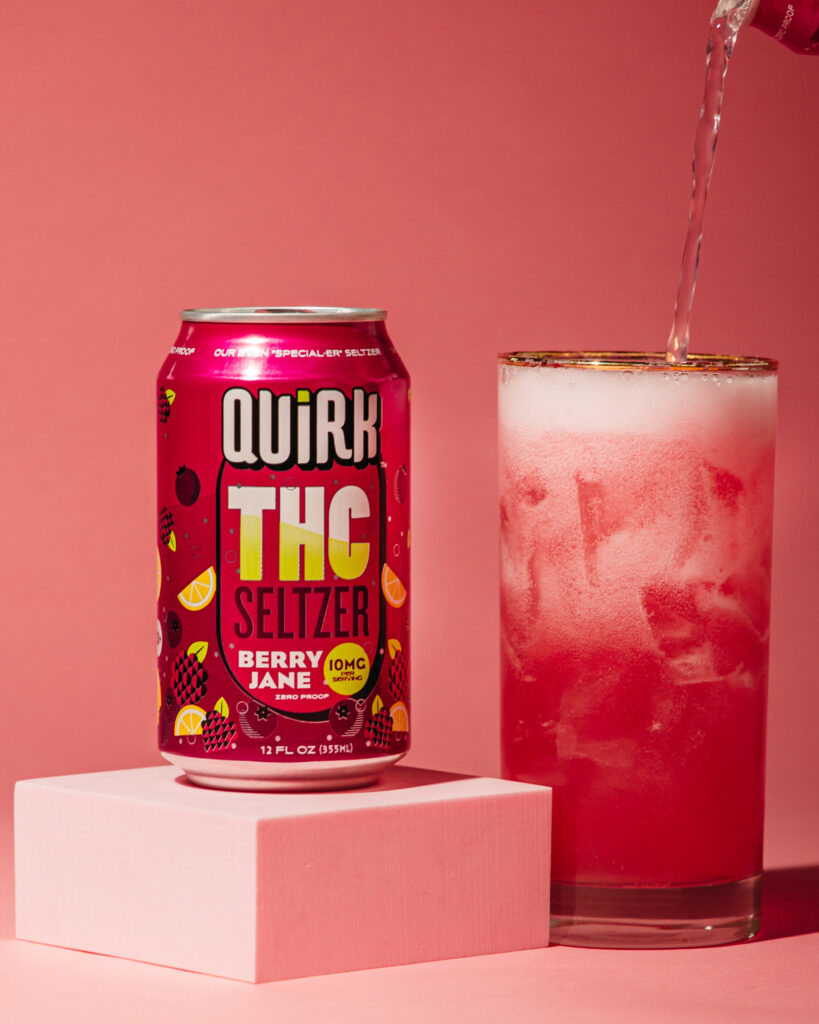 A can of Quirk THC Seltzer Berry Jane flavor sits on a pink block next to a glass of pink THC seltzer with ice, as more seltzer is poured in, all set against a vibrant pink background.