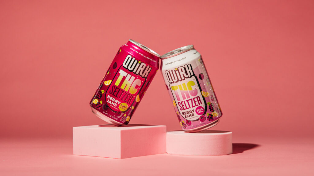Two cans of Quirk THC Seltzer, Berry Lemon flavor, are balanced on pink geometric blocks with a pink background. One THC Seltzer can is dark pink, the other is white, both adorned with vibrant fruit illustrations.