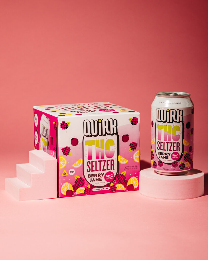 A can and a box of Quirk THC Seltzer, Berry Jane flavor, are displayed on a pink background with small white platforms. The THC Seltzer packaging features vibrant images of berries and citrus fruit slices.