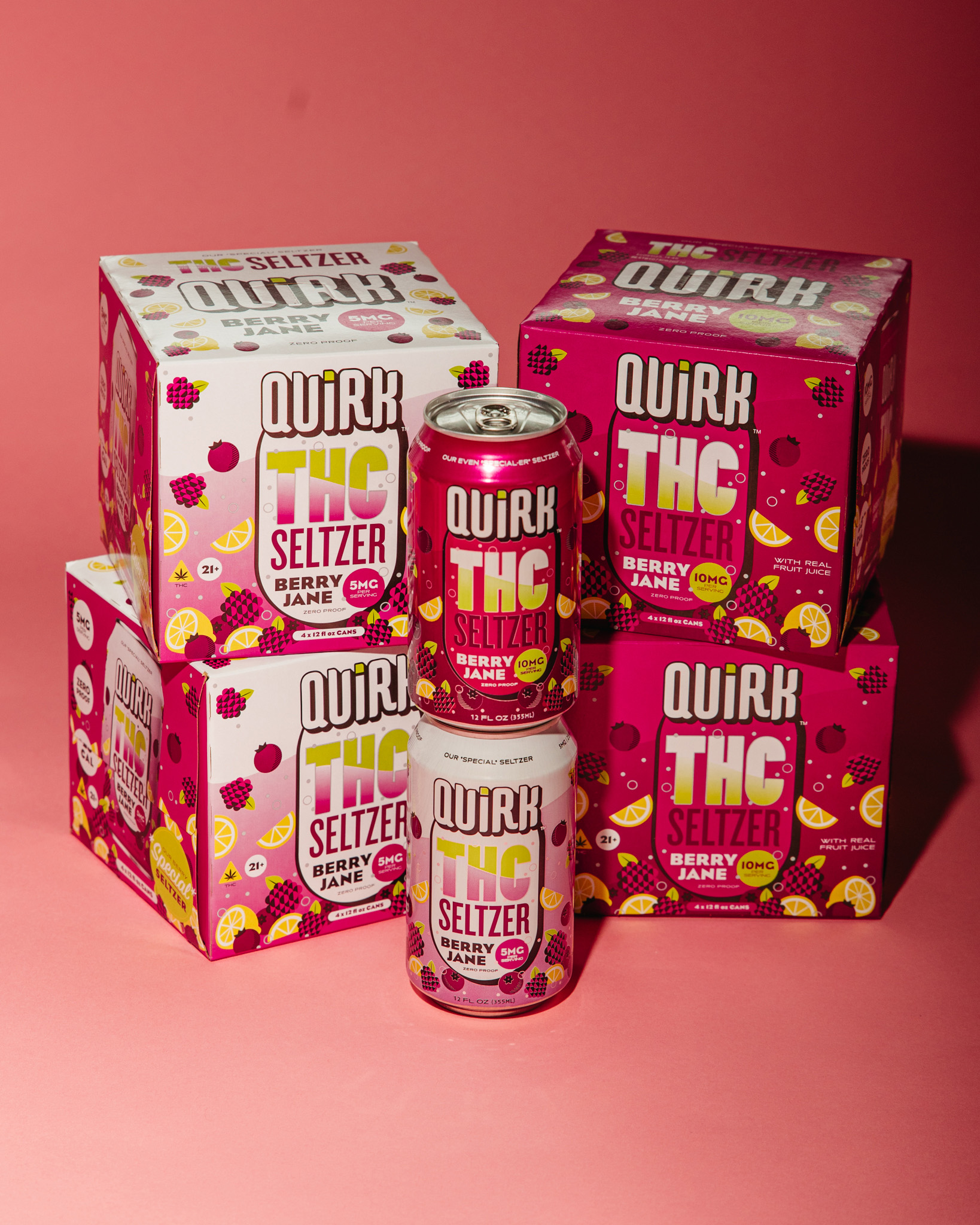 Five boxes and two cans of THC Seltzer Quirk Berry Jane are arranged on a pink surface with a pink background. The colorful packaging features vibrant berry and citrus fruit graphics.