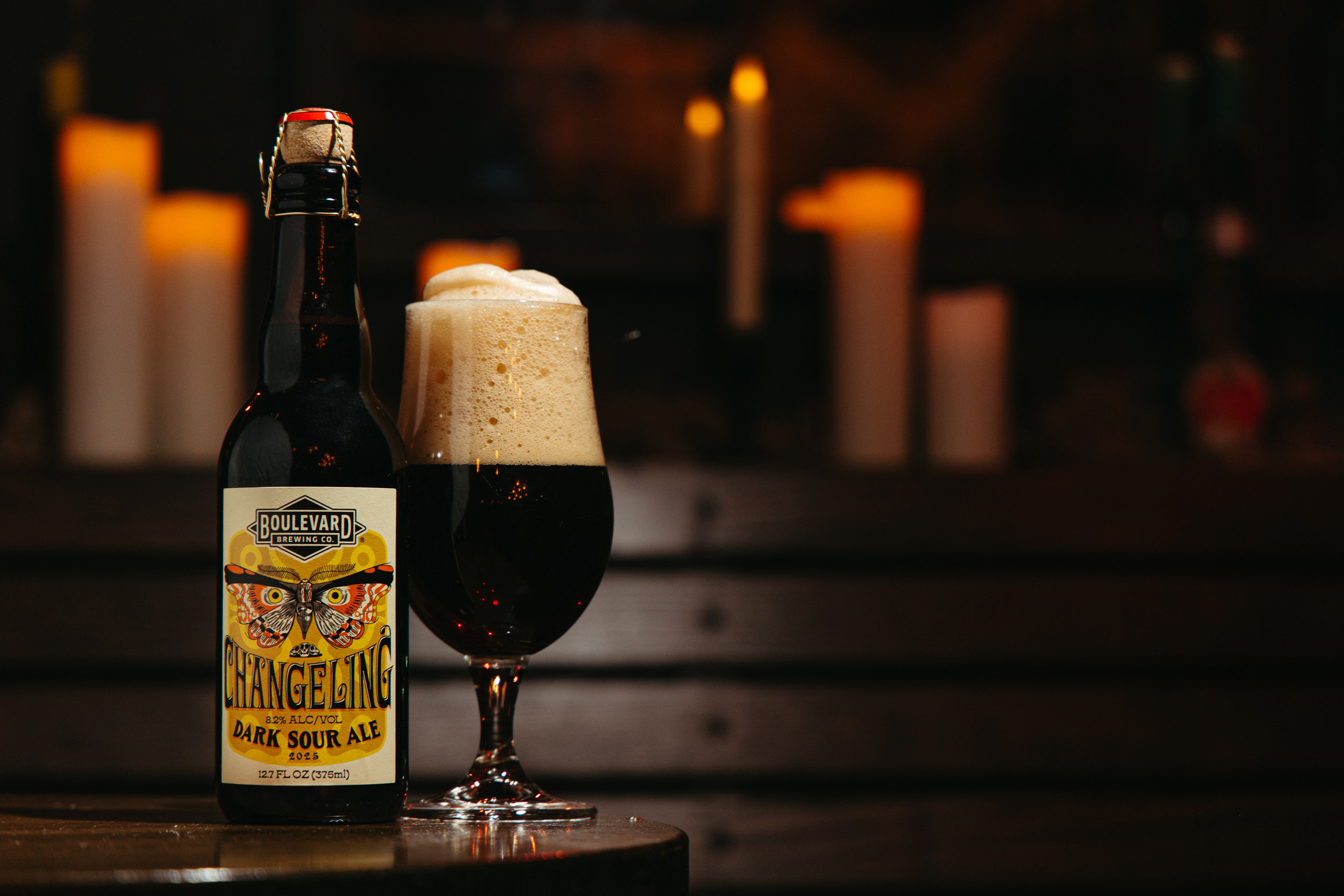 A bottle of Boulevard Brewing Co. Changeling Dark Sour Ale stands next to a glass filled with the ale and a frothy head. Lit candles create a warm, cozy atmosphere in the dimly lit background.