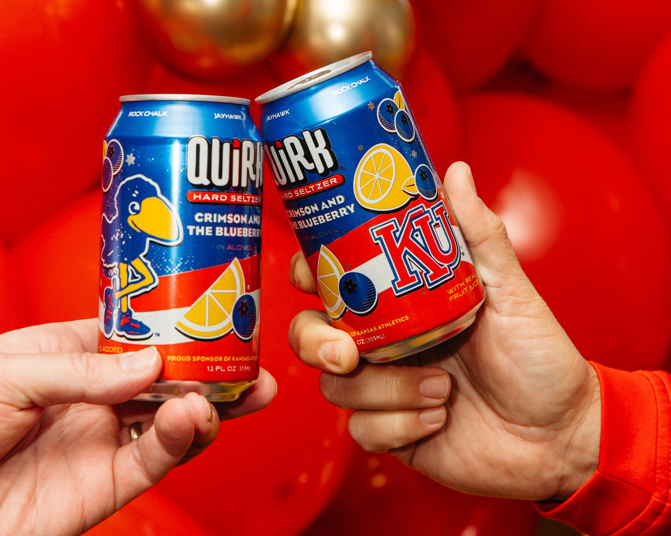 Two hands clink cans of Quirk Hard Seltzer in a crimson and the blueberry design, featuring lemons, blueberries, "KU" letters, and set against a festive backdrop with red balloons.