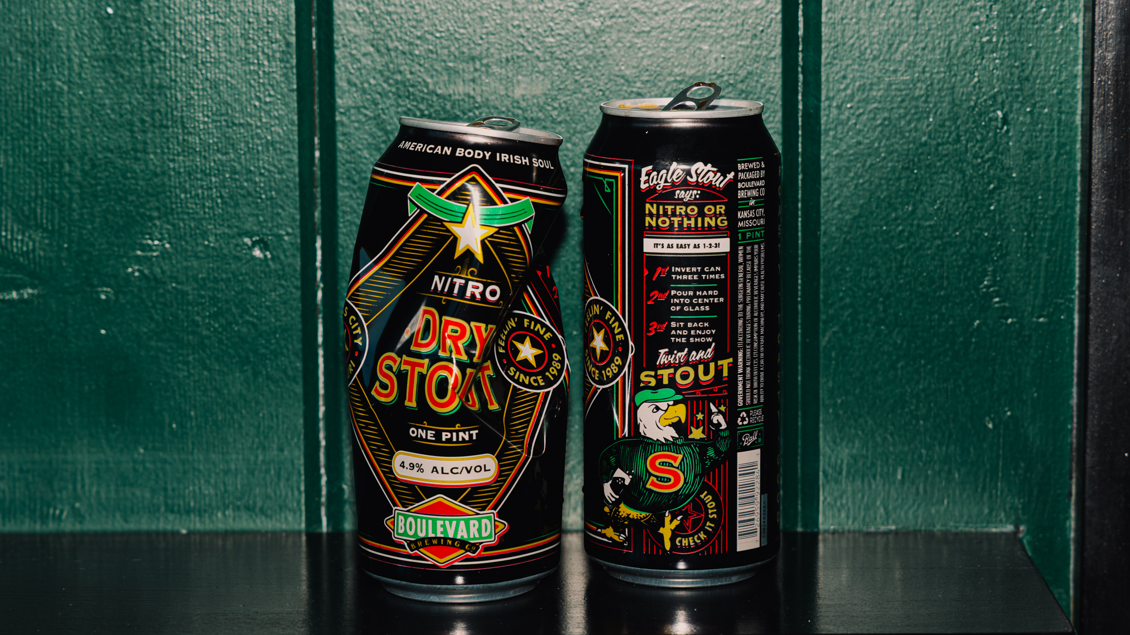 Two tall cans of Boulevard Nitro Dry Stout beer are placed side by side on a dark shelf against a green paneled wall, with one can showing the front label and the other showing the back.