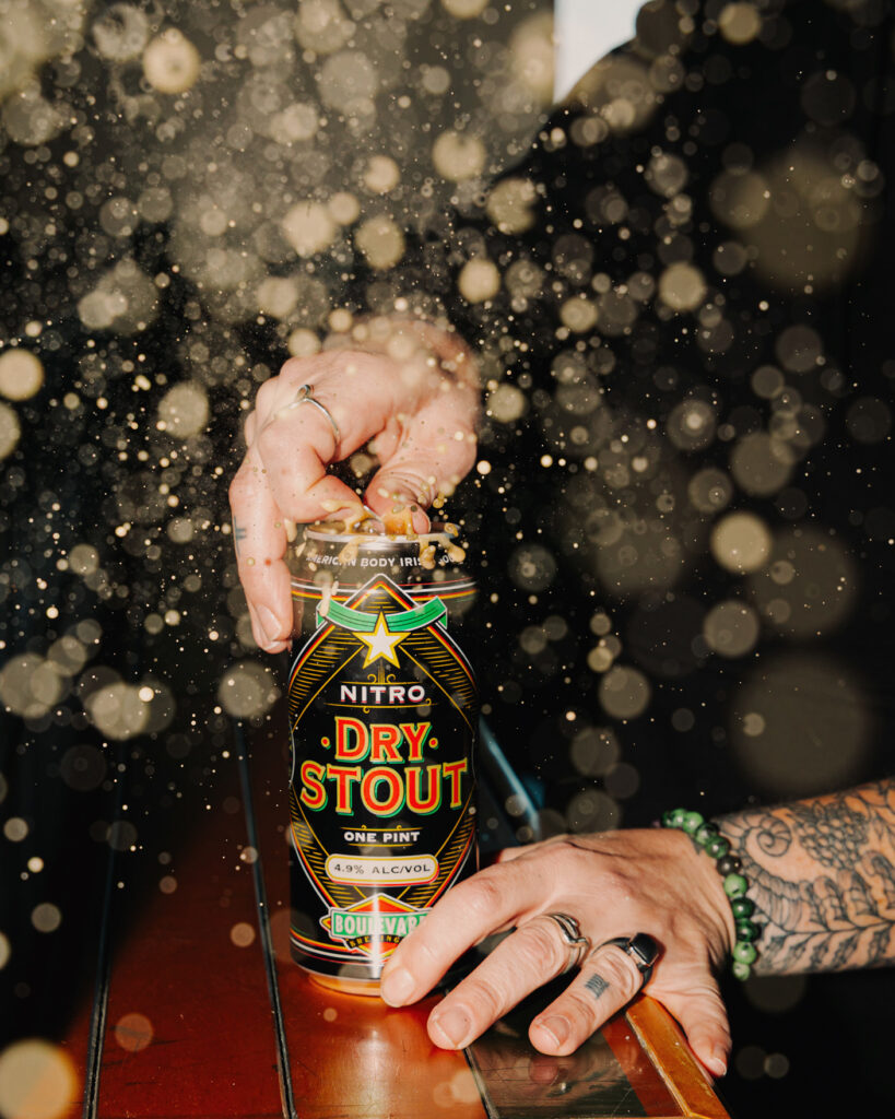 A tattooed person opens a can of Dry Stout beer, with golden bubbles or sparkles floating around, creating a festive atmosphere. The can reads “Nitro Dry Stout, one pint, 4.9% alcohol.”.
