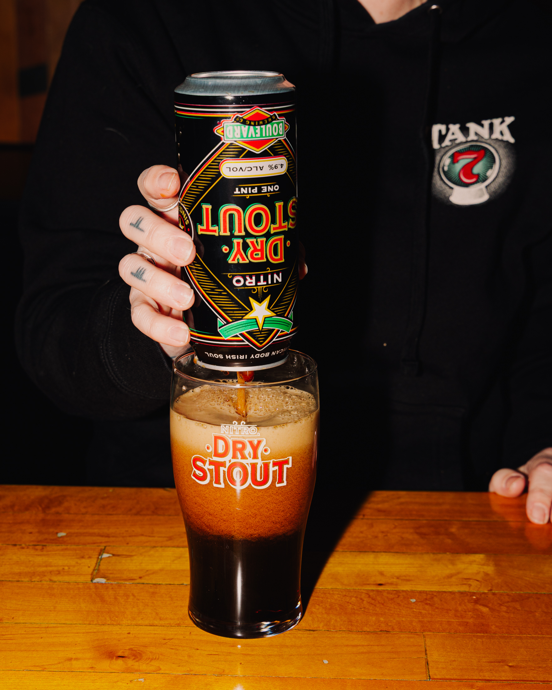 A person pours a can of DRY STOUT Nitro beer upside down into a matching glass on a wooden surface. The hand has tattoos, and the person wears a black hoodie with a circular logo.