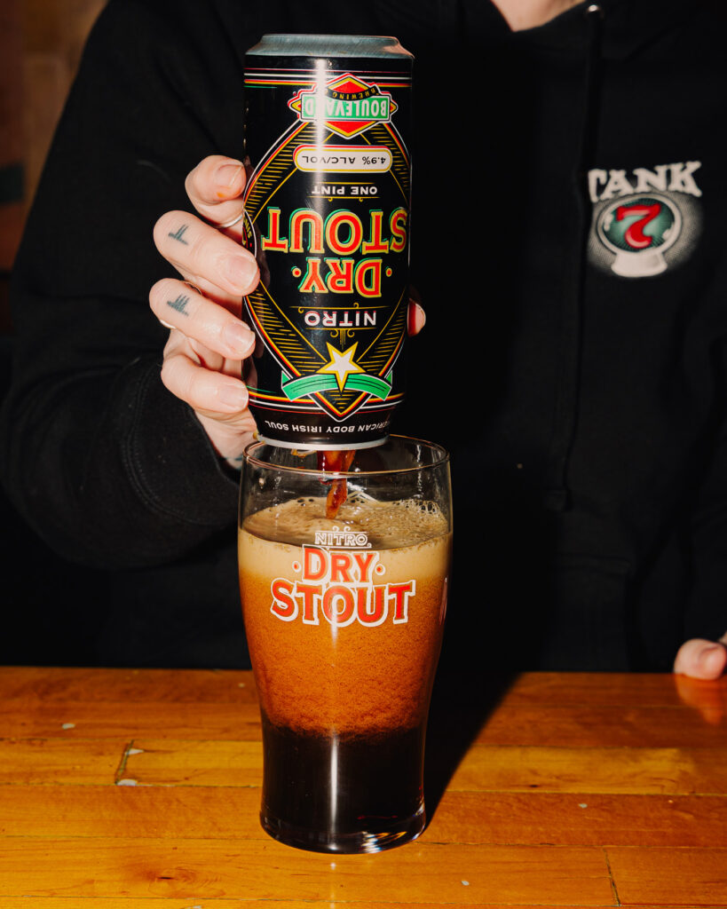 A person pours a can of nitro dry stout beer into a pint glass labeled "Dry Stout" on a wooden table. The beer forms a thick, foamy head as it fills the glass.