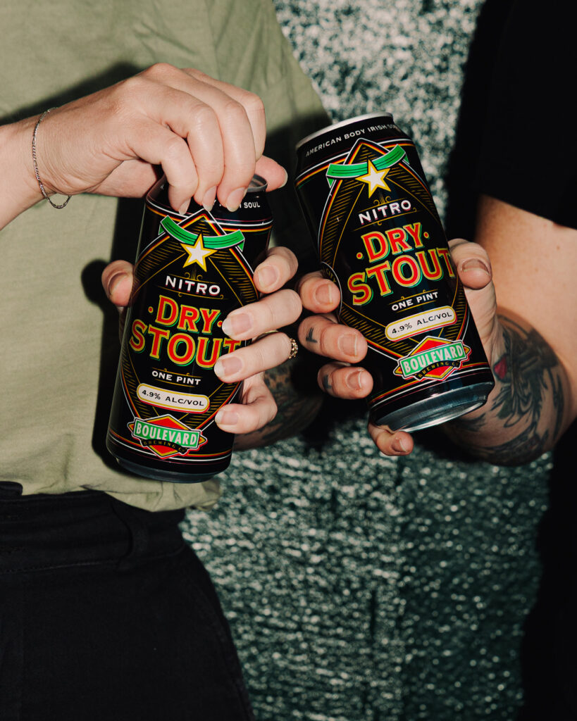 Two people holding Boulevard Nitro Dry Stout cans against a textured background. One person is opening their can, and both have tattoos visible on their arms.