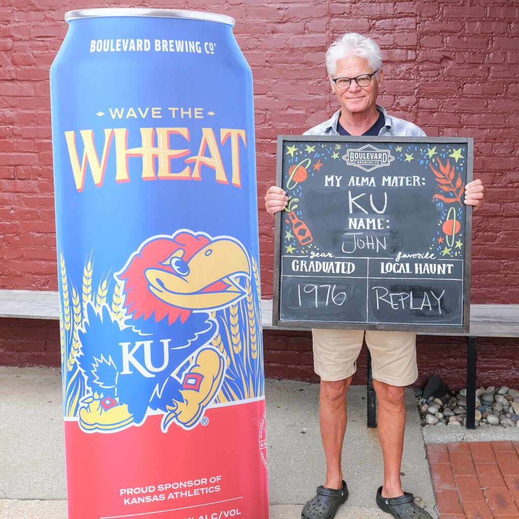 John McDonald holds a chalkboard sign that reads, "My Alma Mater: KU. Name: John. Graduated: 1976. Favorite Local Haunt: Replay." He stands beside a large Boulevard Brewing Co. can.