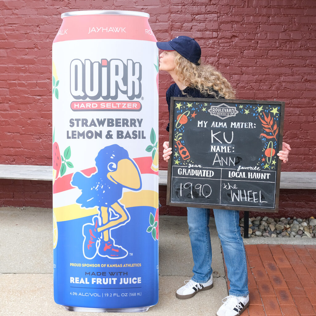 Ann Behner kisses a large can-shaped display of Quirk Strawberry Lemon & Basil Hard Seltzer. Her chalkboard sign features her KU alma mater, name Ann, graduation year 1990, favorite haunt The Wheel.
