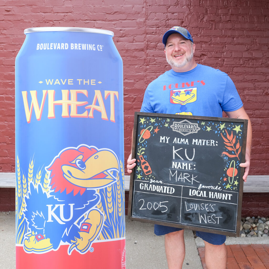 Mark Jackson holds a chalkboard with graduation info, standing next to a large "Wave the Wheat" KU Jayhawk can.