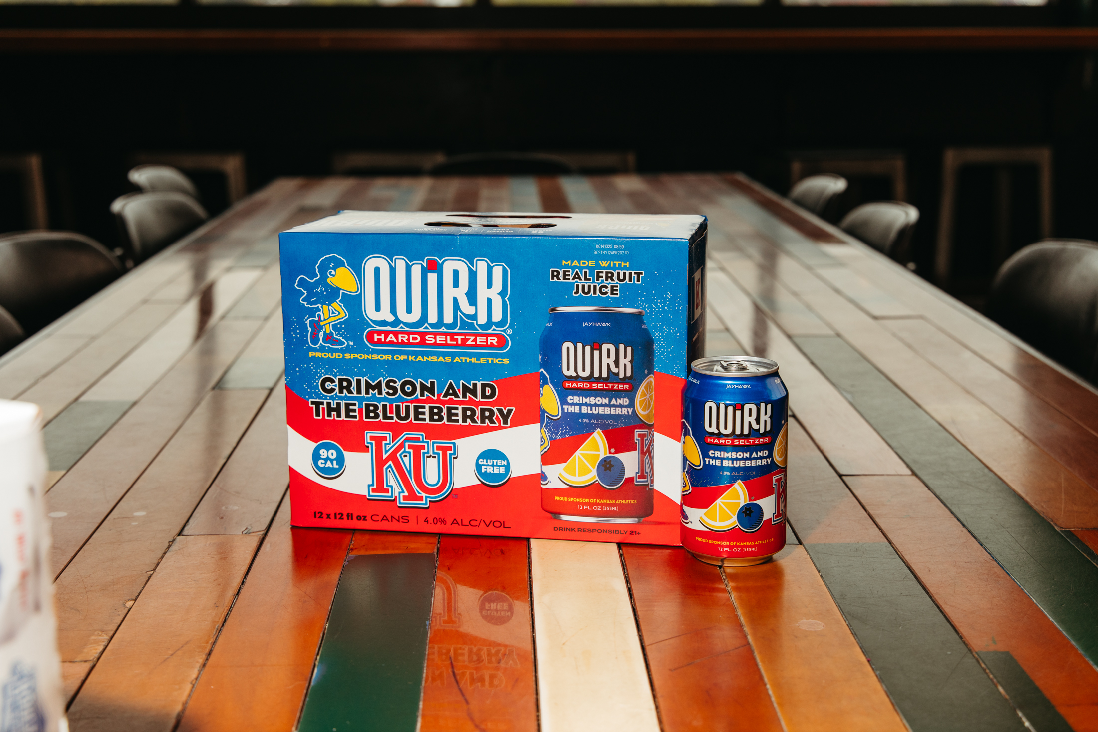 A 12-pack of Quirk Hard Seltzer in the Crimson and the Blueberry flavor sits on a wooden table, featuring KU branding and vibrant images of fruit, with bold blue and red colors on the packaging.