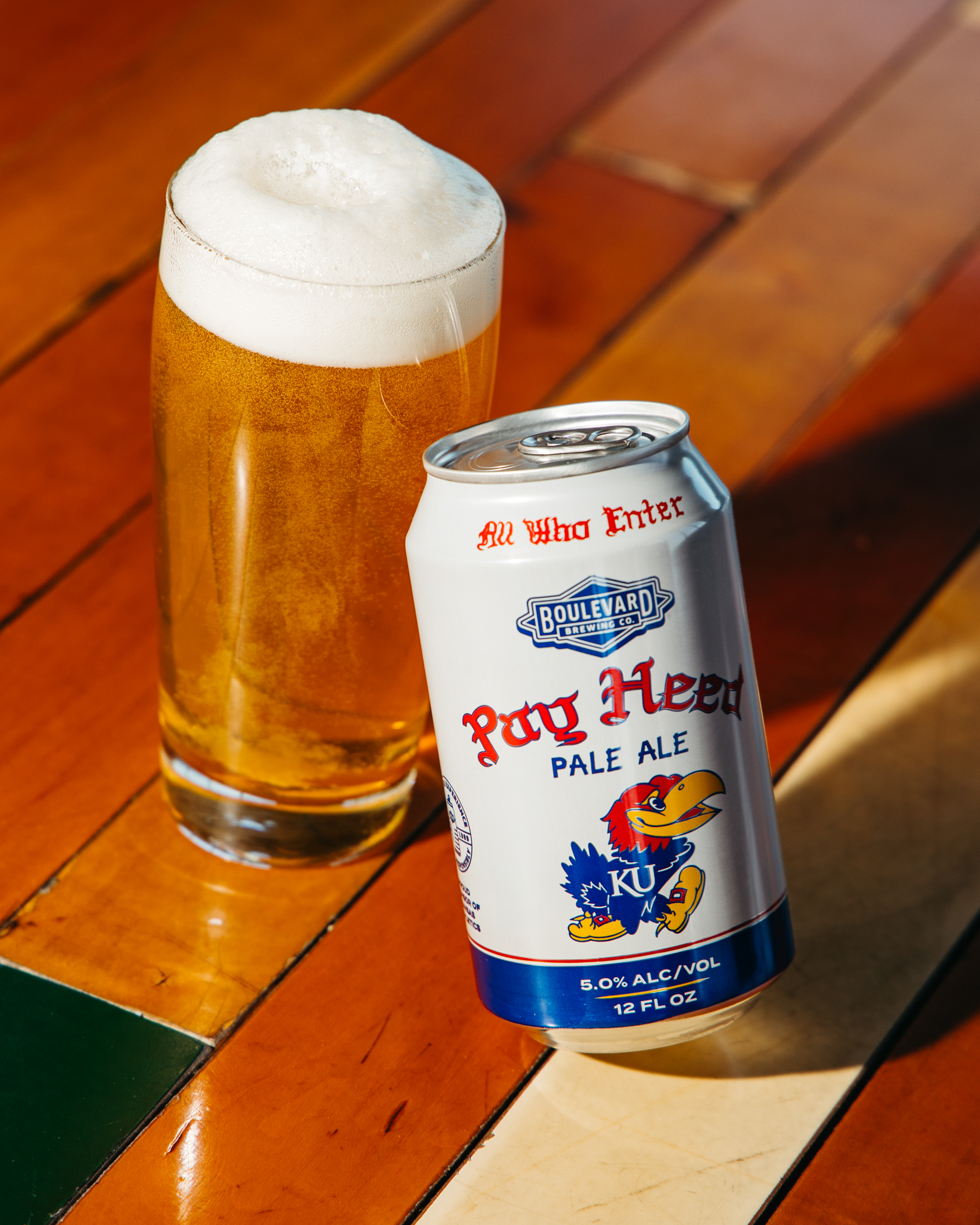 A can of Boulevard Brewing Co. "Pay Heed Pale Ale" with Kansas Jayhawks branding sits next to a tall glass of pale ale on a sunlit wooden surface.
