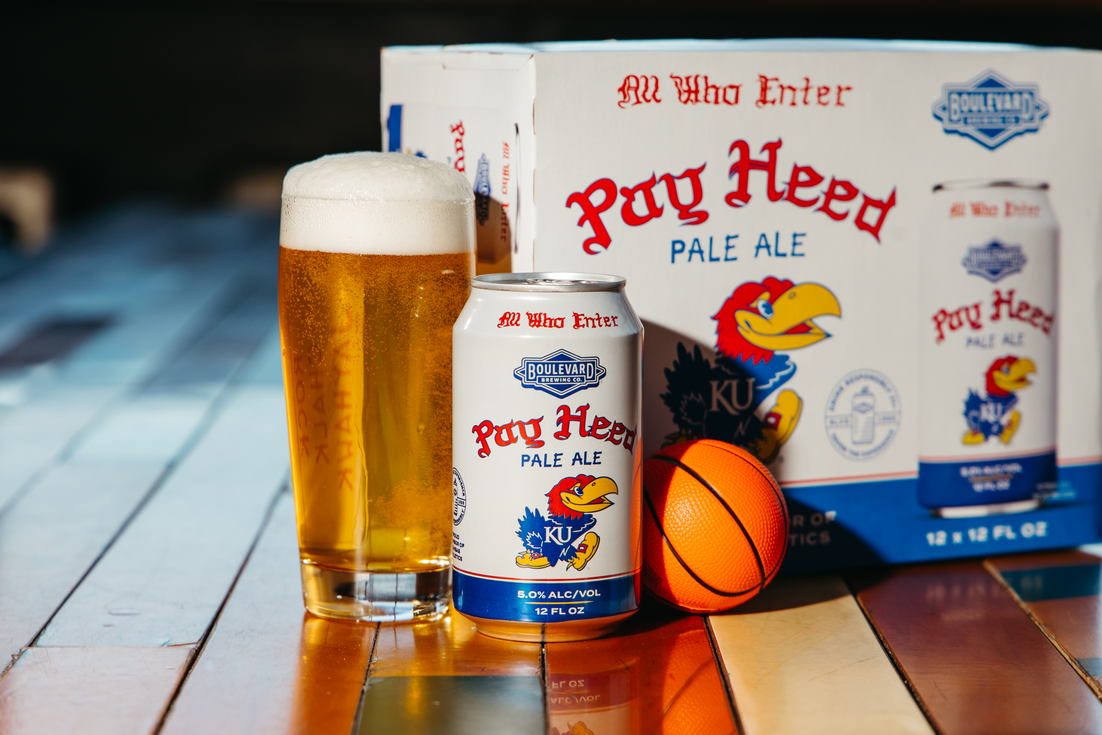A can and a glass of Pay Heed Pale Ale sit on a wooden surface next to a small toy basketball, with a 12-pack box of the beer featuring the Kansas Jayhawks logo in the background.