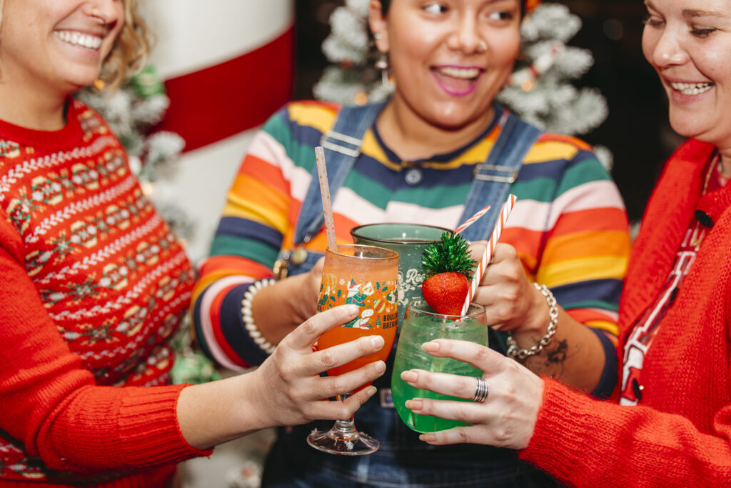 Three people in colorful clothes smile and clink festive holiday drinks together, celebrating Rec Deck the Halls with two decorated Christmas trees blurred in the background.