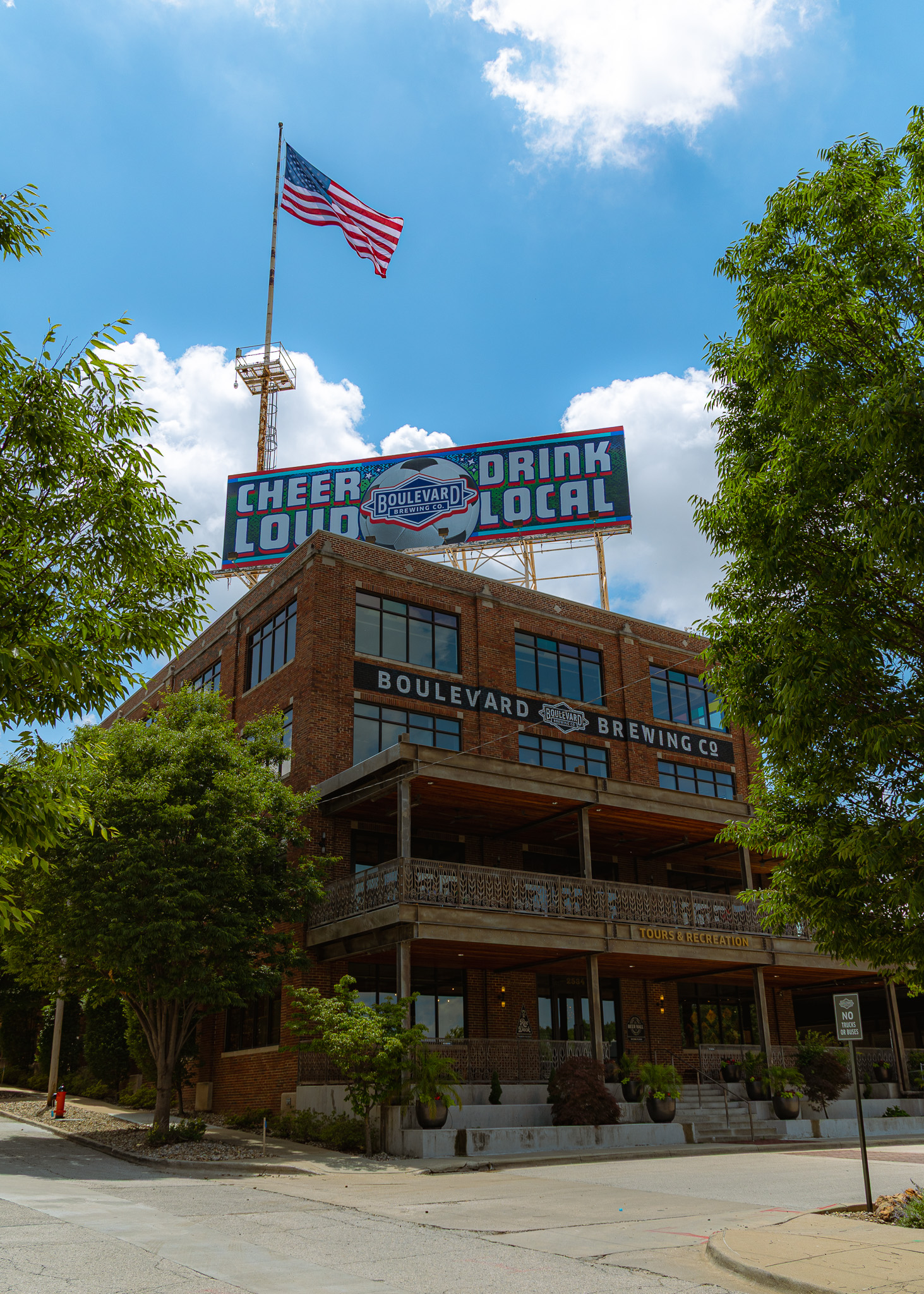 A large brick building with a sign reading "Boulevard Brewing Co" and "Cheers Loud, Drink Local" on the roof, an American flag above, surrounded by trees under a partly cloudy sky.
