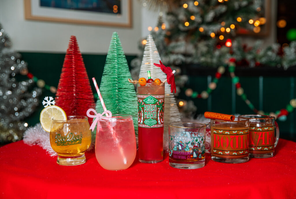 Five festive cocktails on a red tablecloth with holiday decorations set the scene for Rec Deck the Halls, featuring colorful Christmas trees and ornaments. Each drink is garnished with citrus, berries, or cinnamon in decorated glasses.