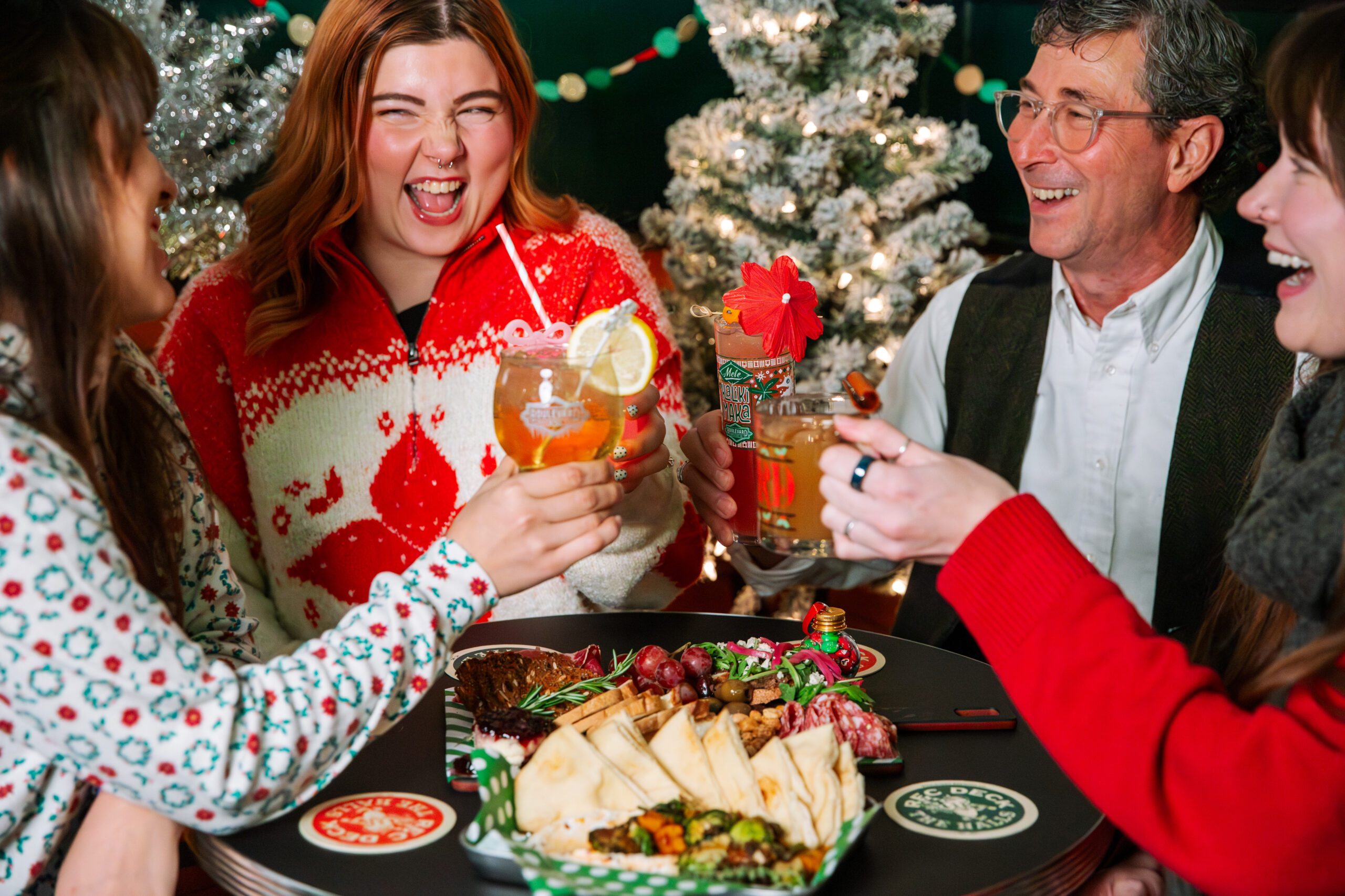 Four people in festive clothing laugh and raise holiday drinks together at a table with assorted appetizers, celebrating the season with Rec Deck the Halls decor—Christmas trees and string lights twinkle in the background.