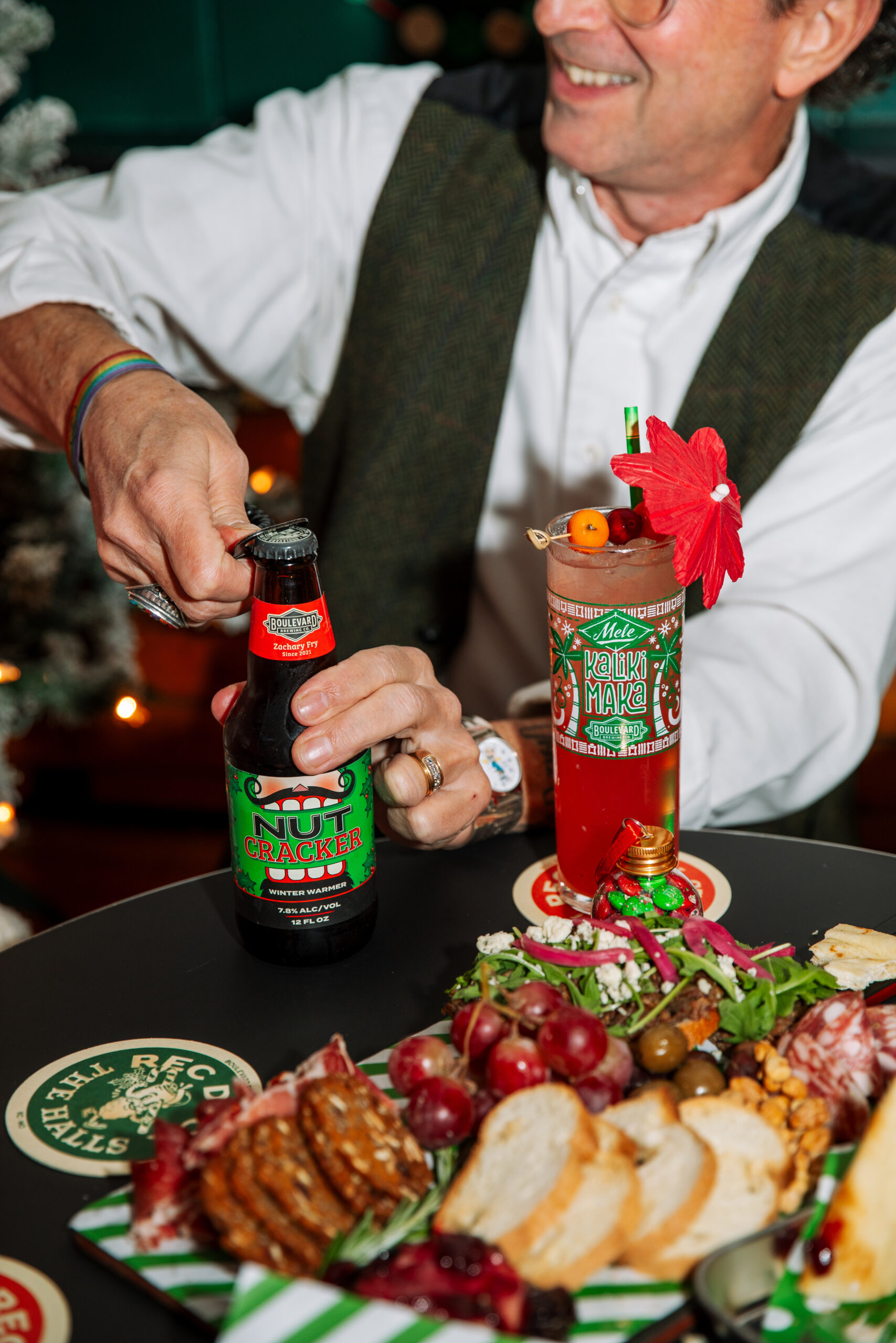 A smiling man opens a bottle of Nut Cracker beer at a festive table for Rec Deck the Halls, surrounded by a holiday cocktail, charcuterie board, bread, grapes, olives, salami, and seasonal décor.