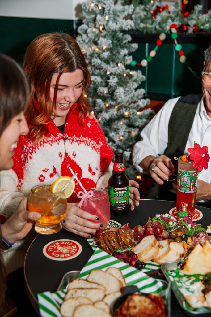Three people sit around a festive table with holiday decorations, drinks, and assorted food. One woman wears a red and white sweater as they rec deck the halls, with a decorated Christmas tree glowing in the background.