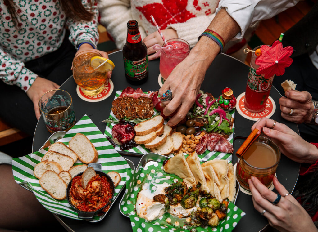 Four people sit around a small round table at Rec Deck the Halls, sharing assorted appetizers like bread, dips, nuts, charcuterie, and cocktails. The scene is festive with colorful drinks and patterned plates on a green-striped paper lining.