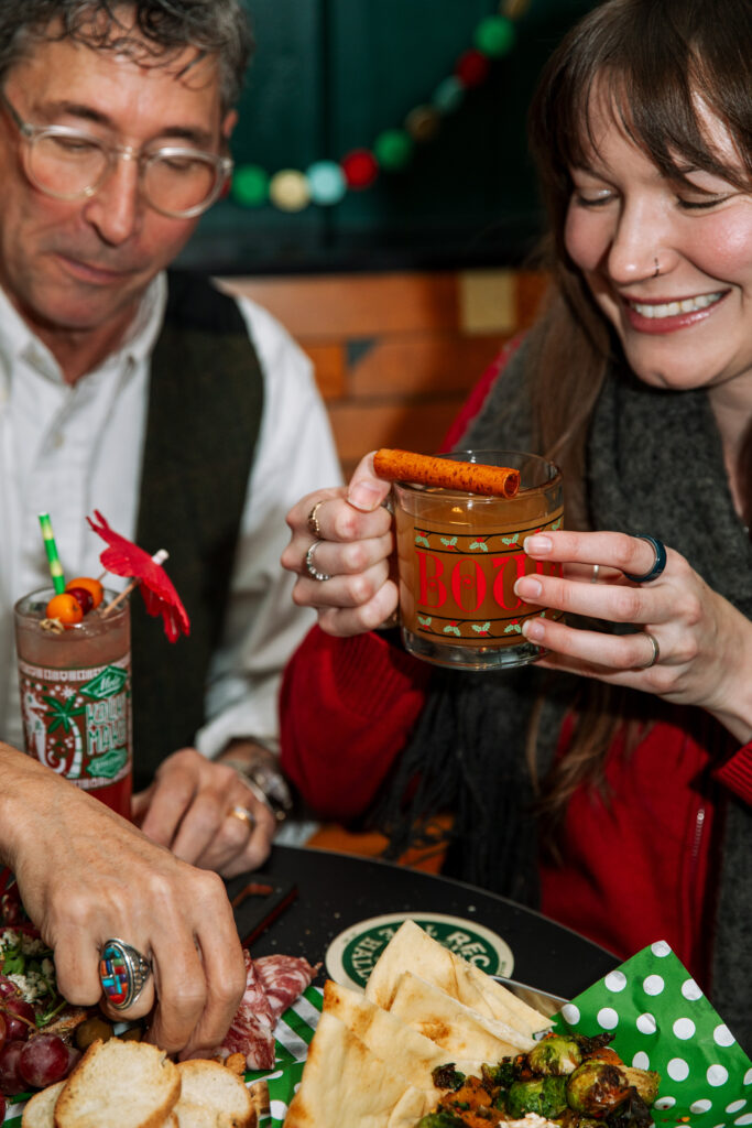 Two people sit at a table with drinks and a plate of food, enjoying a festive meal together at Rec Deck the Halls. One holds a cocktail garnished with cinnamon and an orange slice, while the other reaches for bread.