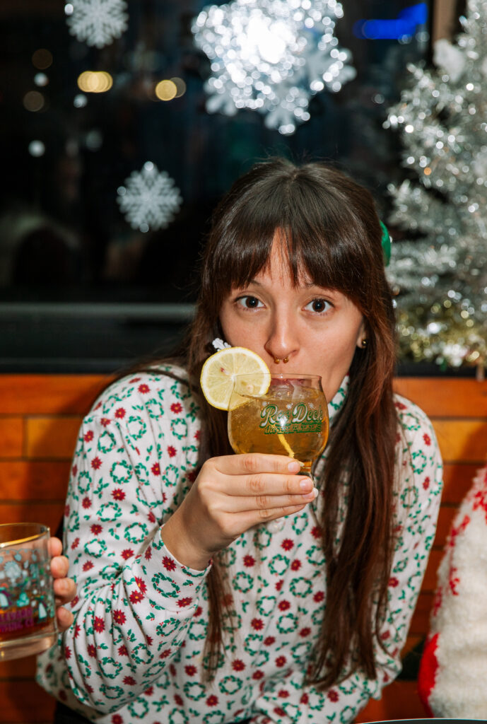 A woman with long brown hair and bangs holds a cocktail garnished with a lemon slice, wearing a festive holiday sweater. Snowflake decorations and a lit Christmas tree set the scene as she gets ready to Rec Deck the Halls.