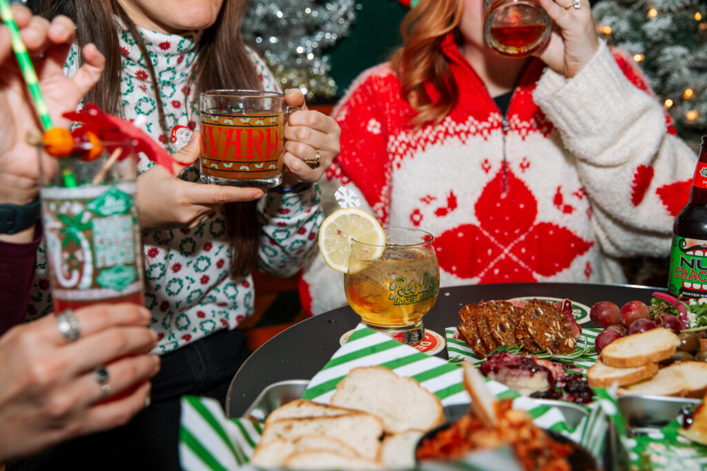 Three people in festive sweaters hold holiday drinks while sitting around a table with assorted snacks, enjoying a cheerful, Rec Deck the Halls-inspired holiday-themed setting.