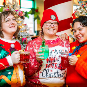 Three people in colorful Christmas sweaters smile and hold festive holiday drinks. Decorated Christmas trees and holiday ornaments are in the background, creating a cheerful, festive atmosphere.